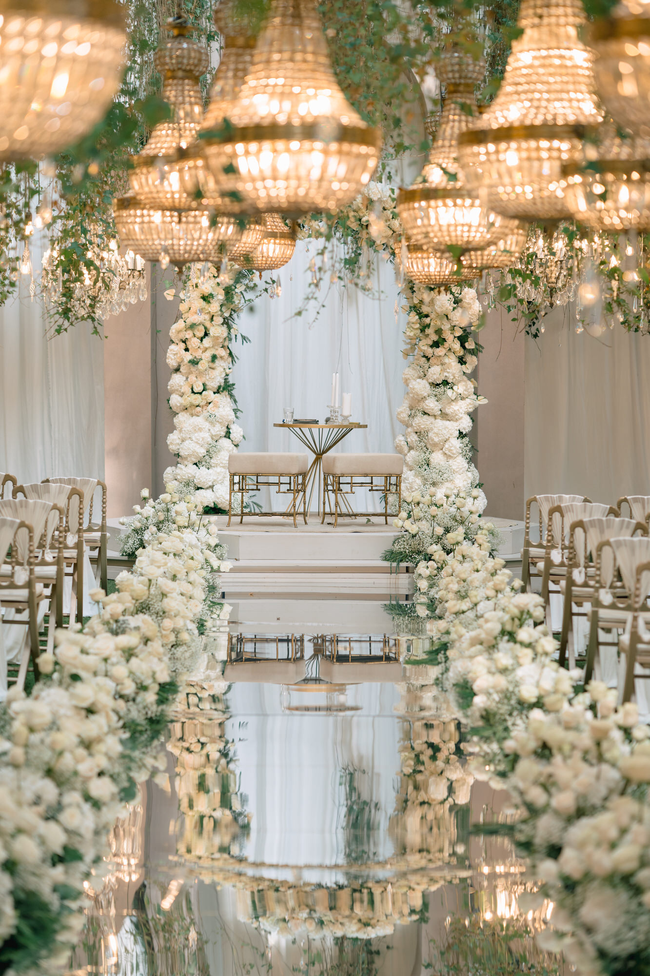 Wide view of the aisle beneath the chandeliers, a floral arch and a gold mirror at its end