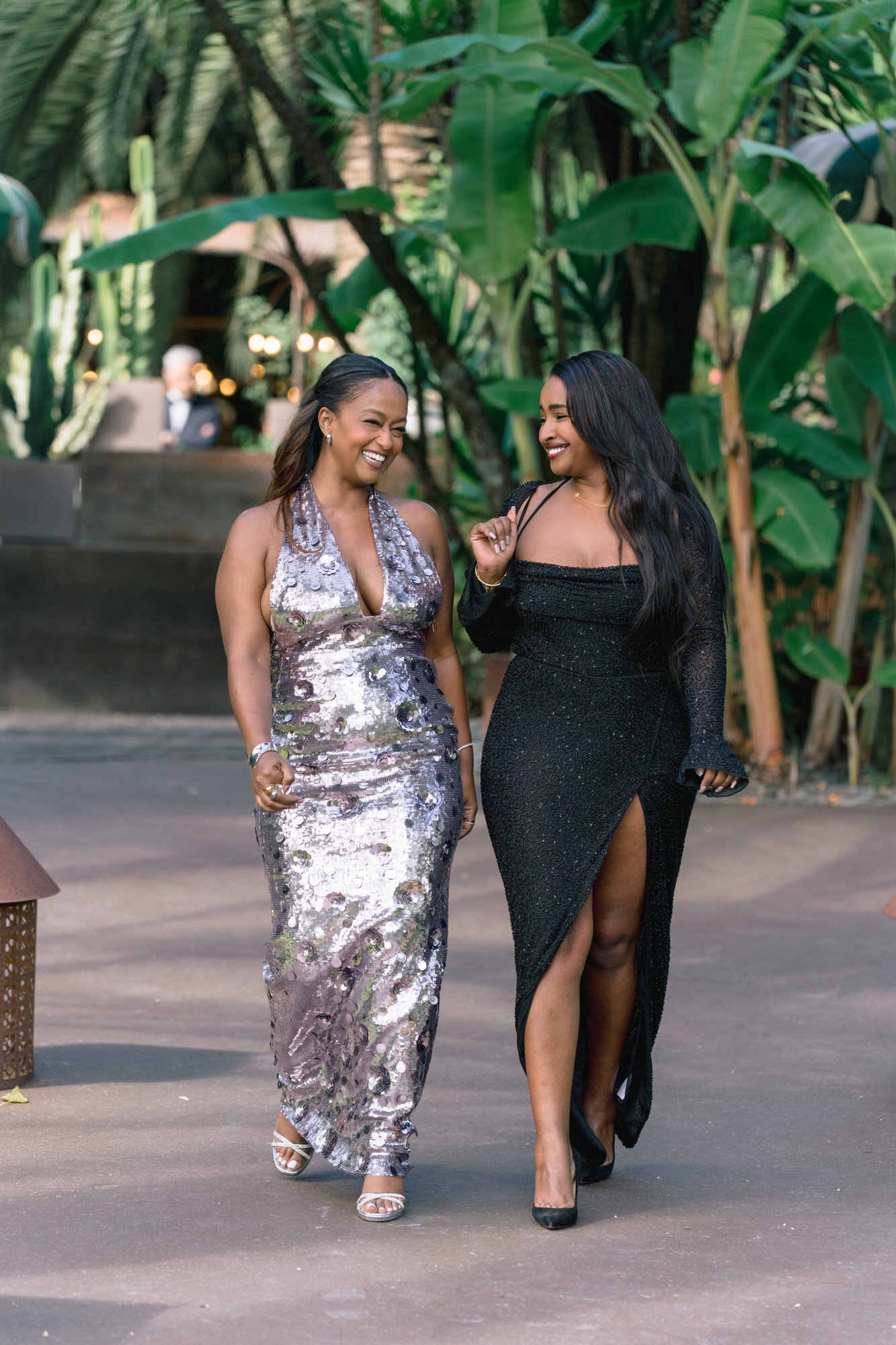 Two guests in a silver sequin dress and a black slit dress walking along the path