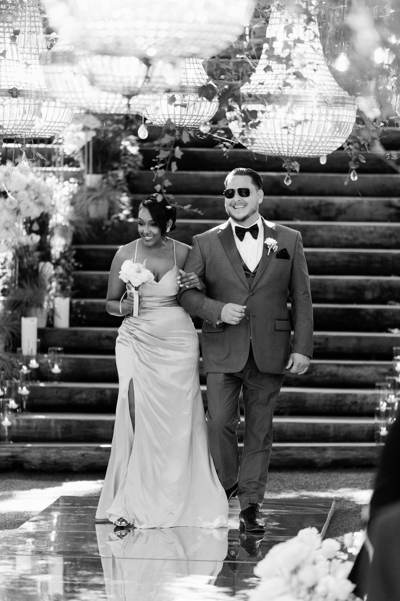 Black-and-white image of the bride on the arm of a man in a dark suit and sunglasses, descending stone steps