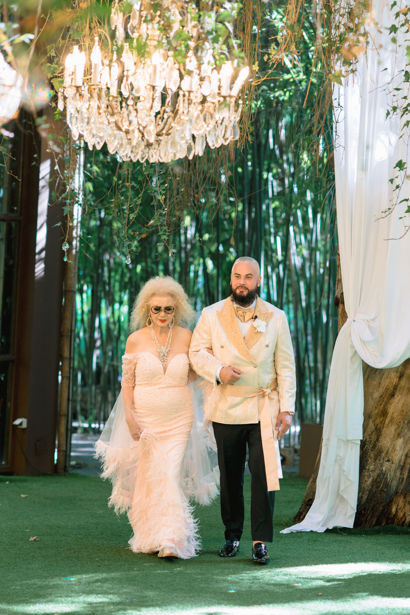 The groom walking down the woodland aisle with a woman in a pale gown beneath a chandelier