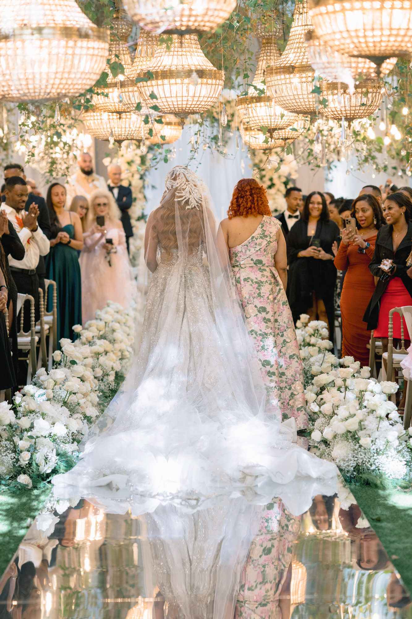 The bride seen from behind as she walks the aisle between guests, her veil trailing over a floral runner