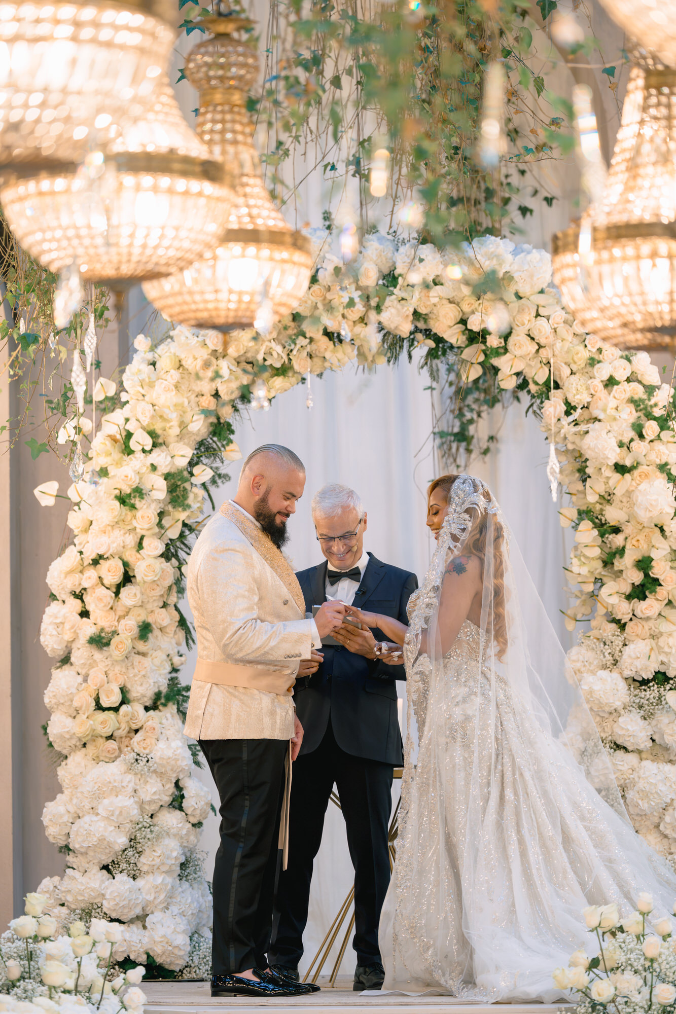 Mahlet and Thomas exchanging vows beneath a white floral arch with celebrant Mariano Sarno, chandeliers above