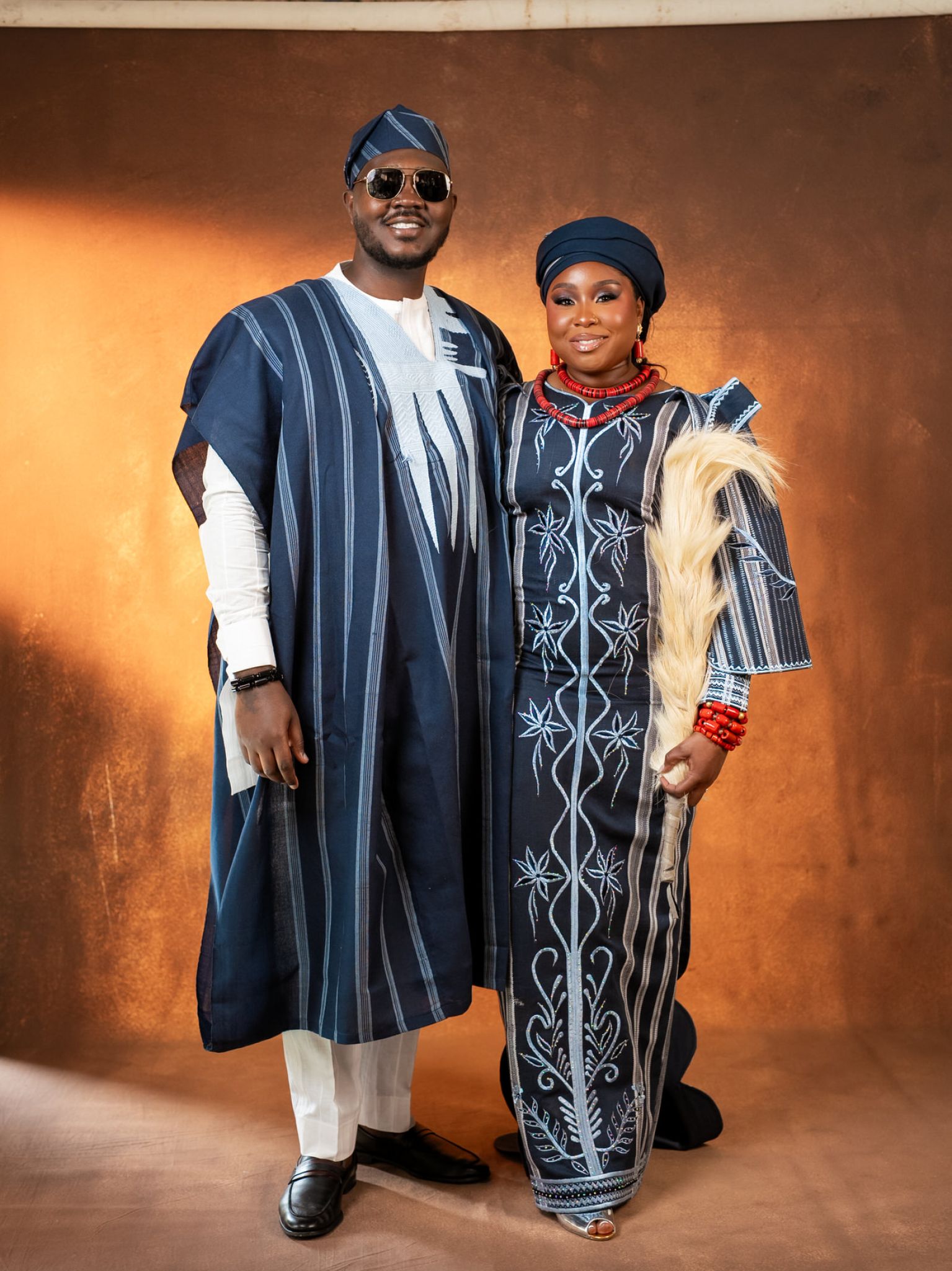 Couple in matching blue traditional outfits smile for the camera indoors against a warm backdrop