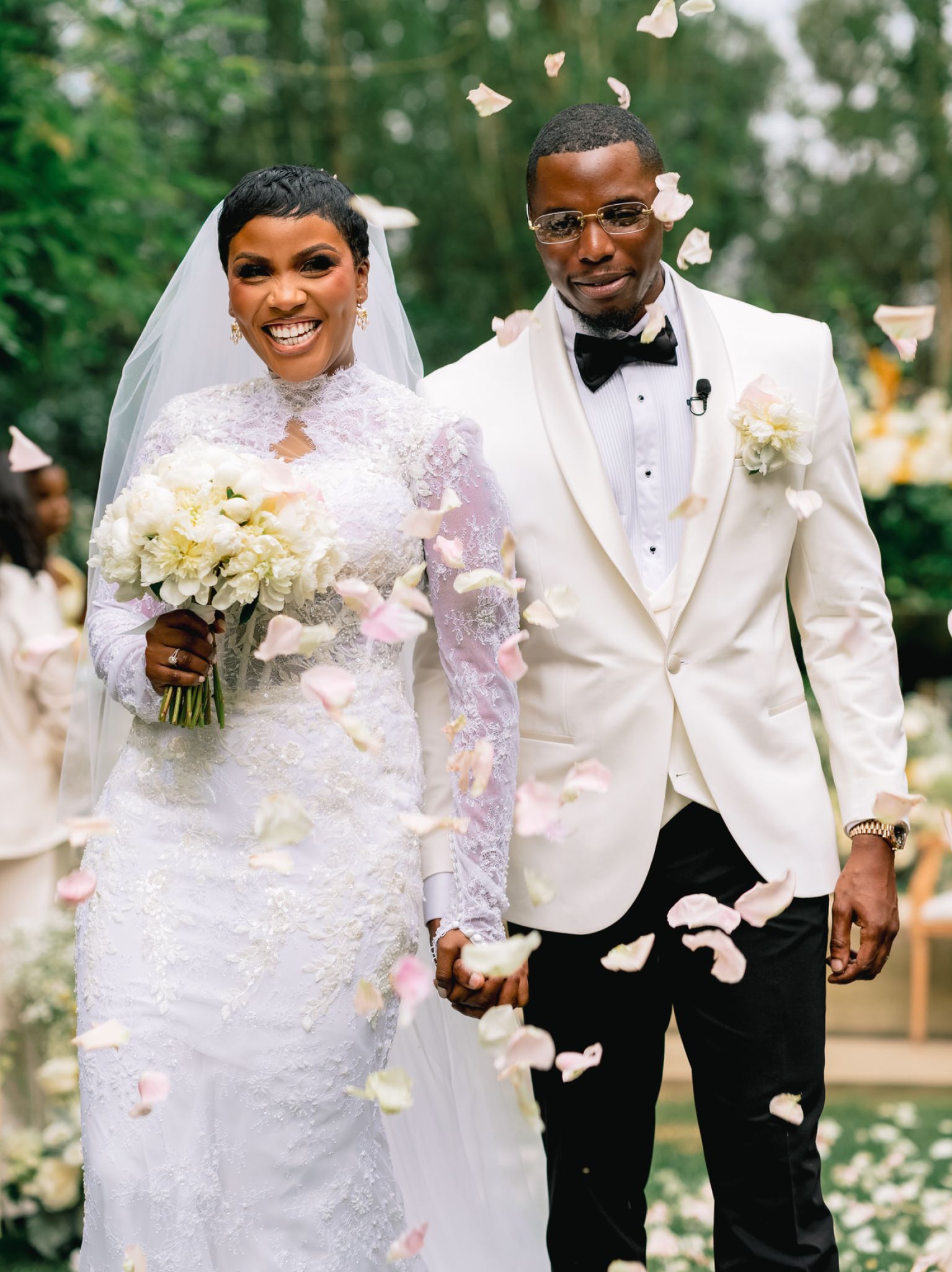 Bride in a white lace wedding dress and veil smiles while holding a bouquet; groom in a white suit with a black bow tie stands beside her as pink petals fall around them.